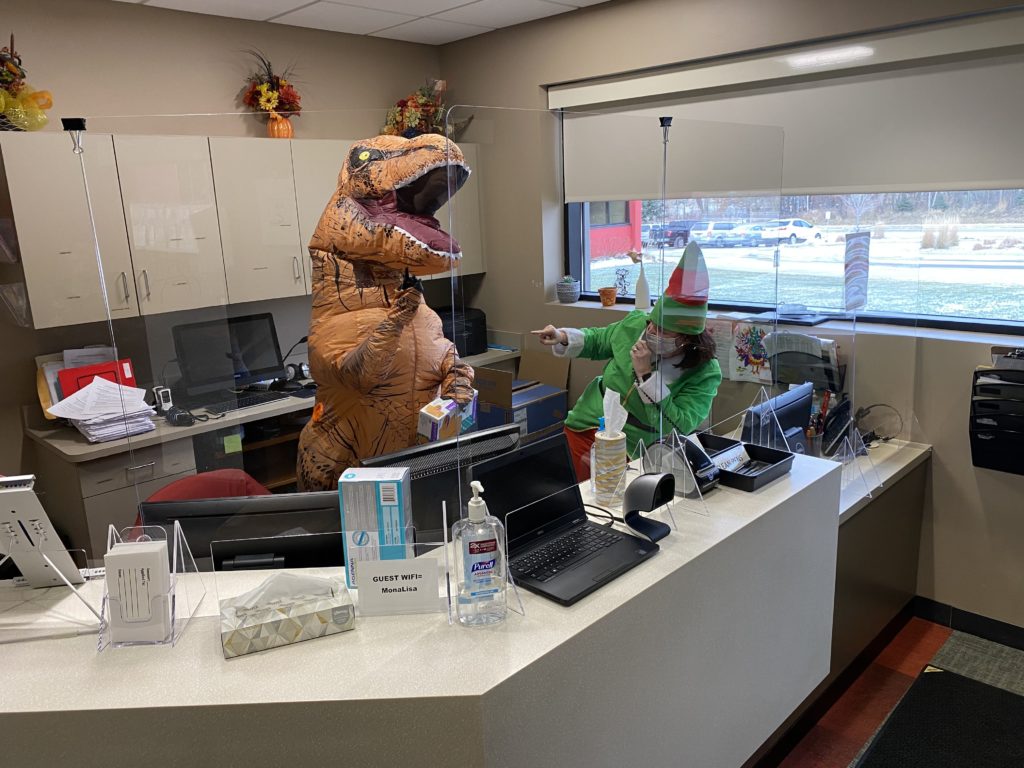 Ms. Zender in a T-rex costume and Ms. Peterson in an elf costume, being playful behind the front desk.
