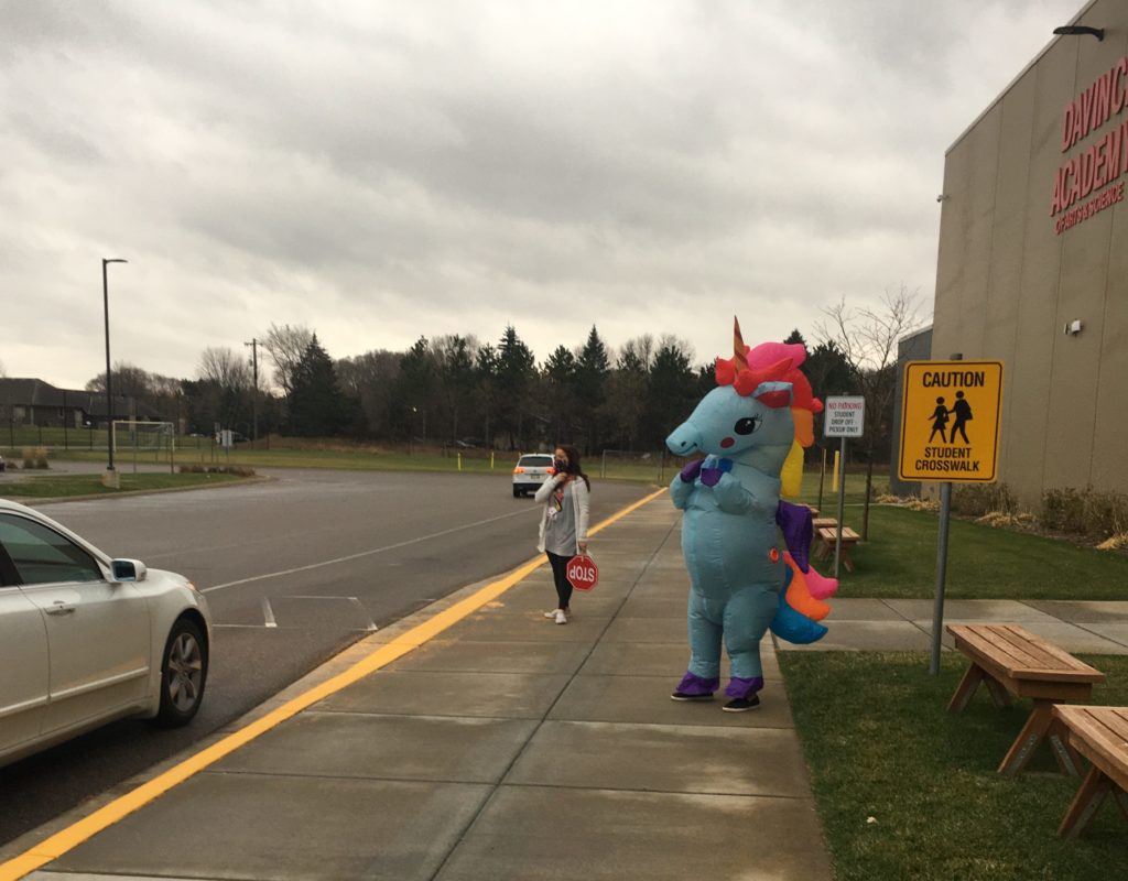 A teacher at morning cross walk duty and another staff member dressed up in a unicorn costume waving to families.