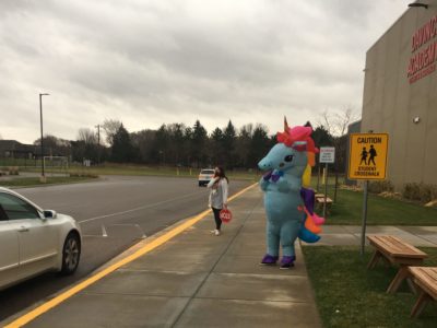 A teacher at morning cross walk duty and another staff member dressed up in a unicorn costume waving to families.